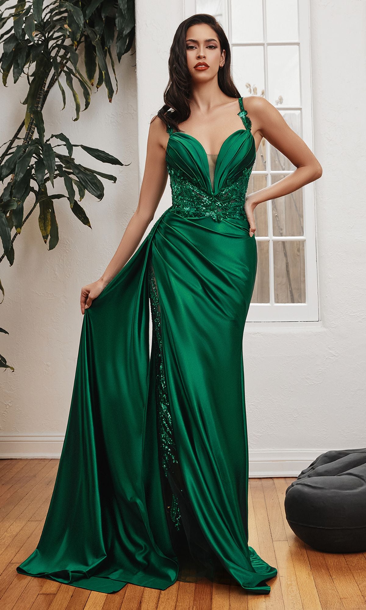 A woman stands indoors in the Long Satin Formal Dress with Side Sash CDS417, an emerald green floor-length gown featuring a deep neckline, lace details, a high slit, and holding up one side of the elegant satin dress.